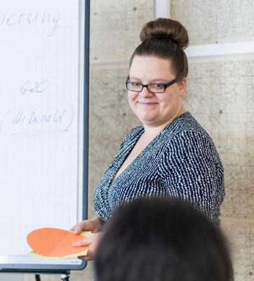 Eine Frau mit hochgesteckten Haaren und Brille steht neben einem Whiteboard und hält einen Vortrag.