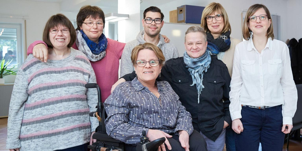Sechs Frauen und ein Mann stehen für Gruppenbild zusammen. Die Frau in der Mitte sitzt in einem E-Rolli.