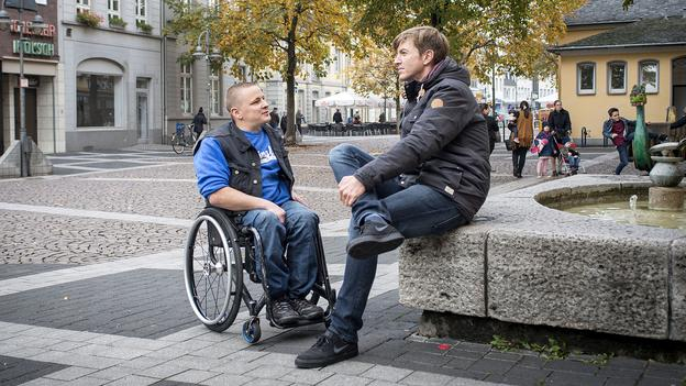 ein rollstuhlfahrer und ein junger mann sitzend auf einem brunnenmauer in einer fußgängerzone