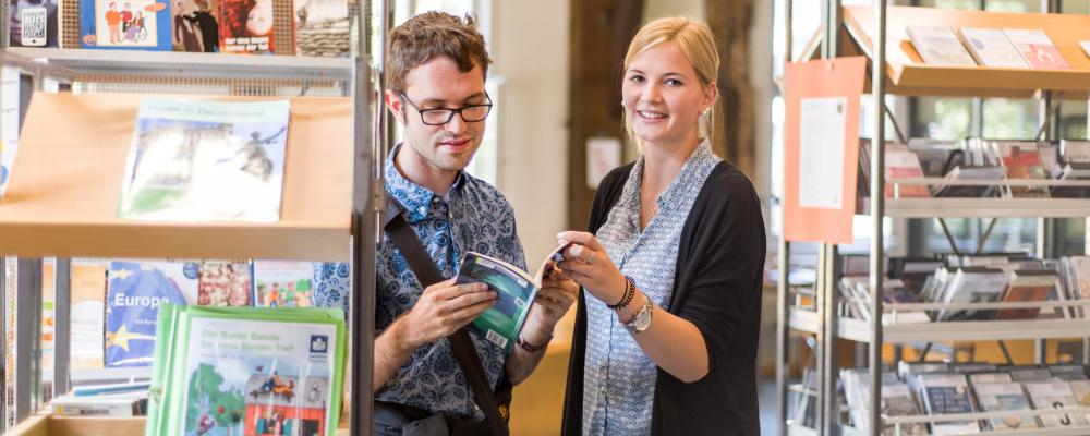 Ein Mann und eine Frau stehen neben Bücherregalen in denen Bücher in Leichter Sprache liegen