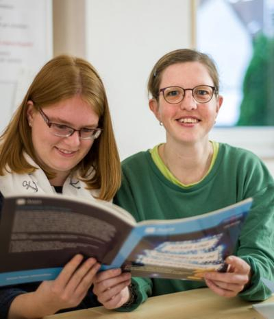 Zwei Frauen sitzen an einem Schreibtisch und halten ein Buch in den Händen, die linke Frau liest, die rechte Frau lächelt in die Kamera.