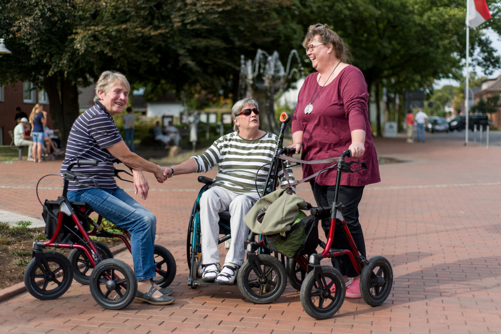 Drei Frauen mit unterschiedlichen Mobilitätseinschränkungen  unterhalten sich  auf einem Platz. 