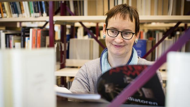 Agnes steht vor einem Bücherregal und liest eine Zeitschrift.