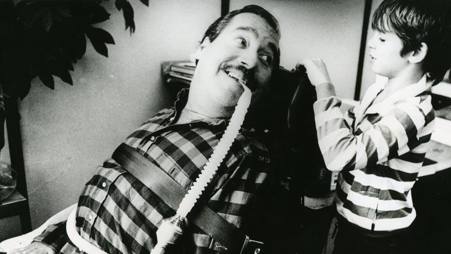 Ein Mann mit Beatmungsschlauch im Rollstuhl schaut lächelnd zu einem Jungen 