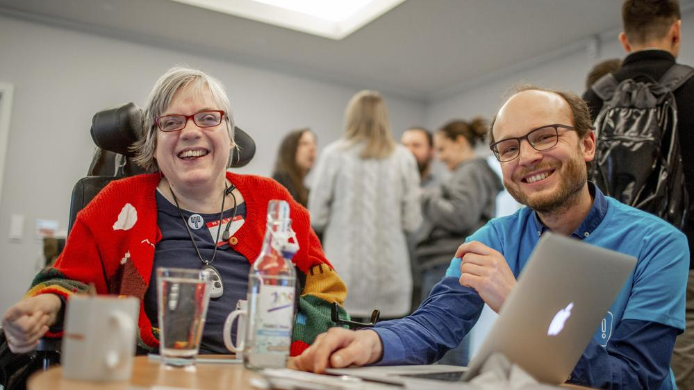 Eine Frau im Rollstuhl und ein Mann lachen in die Kamera.