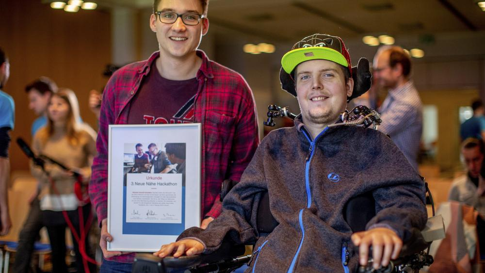 Zwei Männer, einer davon im Rollstuhl, lächeln in die Kamera.