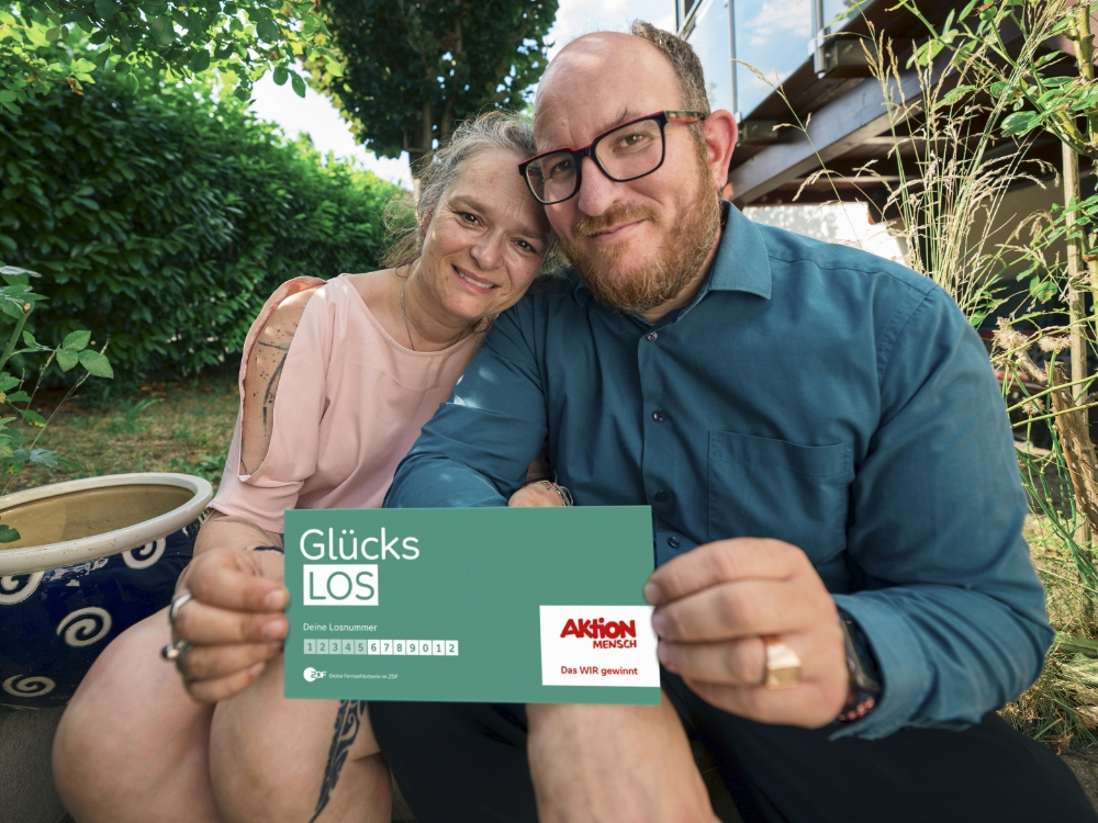 Ein Paar sitzt zusammen im Freien, sie lächeln freundlich. Sie halten ein Glücks-Los der Aktion Mensch in der Hand. Die Atmosphäre wirkt entspannt und freundlich.