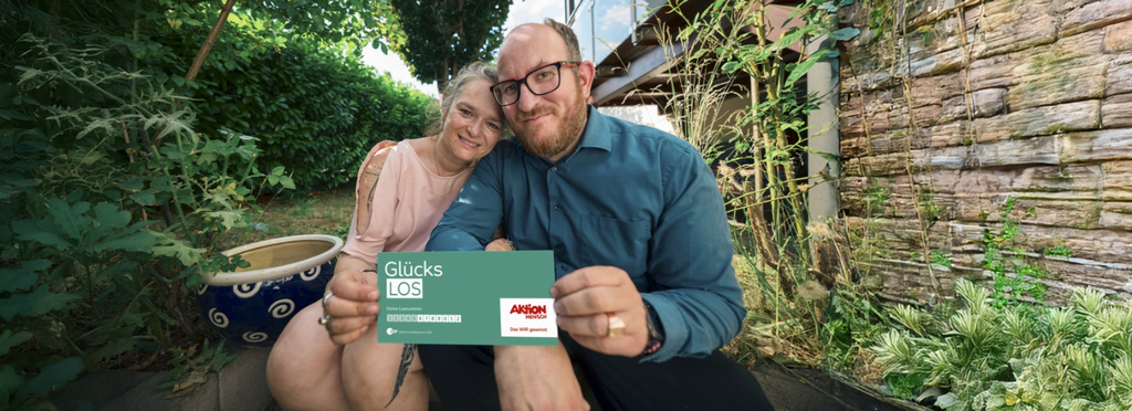 Ein Paar sitzt zusammen im Freien, sie lächeln freundlich. Sie halten ein Glücks-Los der Aktion Mensch in der Hand. Die Atmosphäre wirkt entspannt und freundlich.