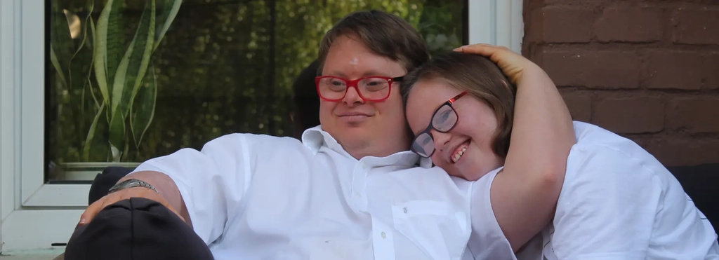 Ein junges Pärchen - beide mit Downsyndrom - sitzen auf dem Sofa. Er hat seinen Arm um sie gelegt und sie kuschelt sich an ihn an.