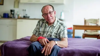 Älterer Mann mit Brille in Jeans und kariretem Hemd sitzt auf dem blau bezogenen Bett seines Appartements und lächelt in die Kamera