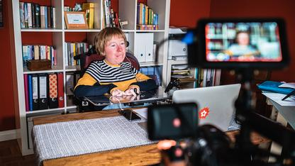 Eine Frau im Rollstuhl sitzt an einem Tisch, im Vordergrund ist eine Kamera zu sehen, die sie filmt.