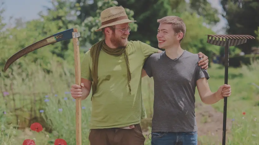 In einem Garten stehen ein älterer und ein jüngerer Mann nebeneinander. Sie haben die Arme umeinander gelegt und haben Gartengeräte in der Hand.