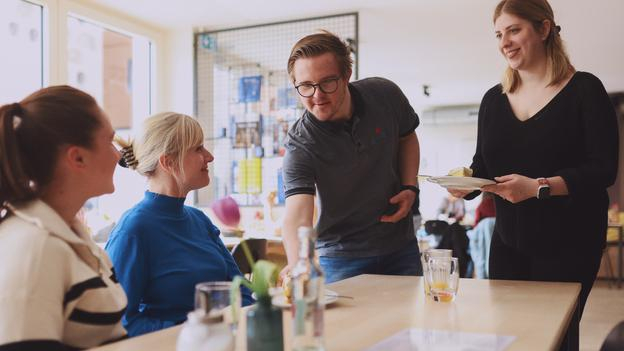 Zwei Frauen sitzen an einem Holztisch. Ein Mann mit Down-Syndrom und eine junge Frau servieren jeder einen Teller mit Kuchen.