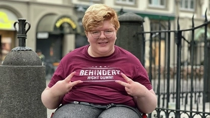 Eine junge Frau im Rollstuhl. Sie trägt ein T-Shirt mit der Aufschrift: Behindert - Nicht Dumm!
