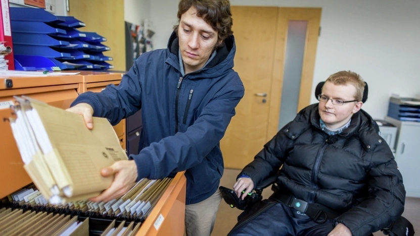 Ein Mann steht neben einem anderen jungen Mann im Rollstuhl und holt Akten aus einem Schrank.