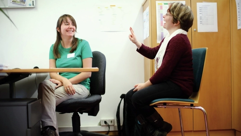 Zwei Frauen sitzen um einen Schreibtisch. Eine der beiden gebärdet.