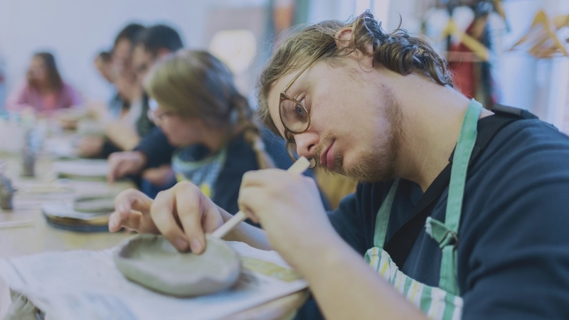 An einem Werktisch in einem Atelier sitzen mehrere junge Erwachsene und arbeiten mit Ton. Der junge Mann vorne im Bild ist scharf zu sehen. die dahinter sind verschwommen. Konzentriert zieht der junge Mann mit blonden gewellten Haaren und Brille den Rand einer kleinen Schale hoch, die er aus dem Ton gerade formt.