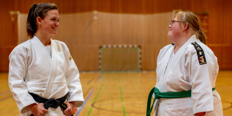 In einer Sporthalle stehen zwei Frauen nebeneinander. Sie tragen weiße Judo-Anzüge und schauen sich freundschaftlich an. Eine der beiden Frauen hat eine Behinderung.