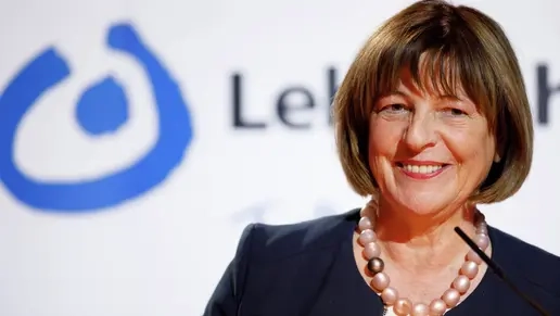 Eine Frau, Ende 60, mit braunen Haaren im Pagenkopfschnitt, geschminkten Lippen einer Kette mit dicken Kugeln und blauem Kostüm lächelt leicht an der Kamera vorbei. Im Hintergrund etwas verschwommen das Logo der Lebenshilfe.