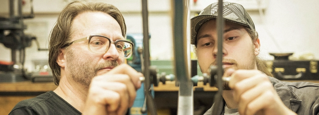 Ein jüngerer und ein mittelalter Mann stehen gemeinsam an einem Werktisch einer Fahrradwerkstatt und bedienen ein Werkzeug.
