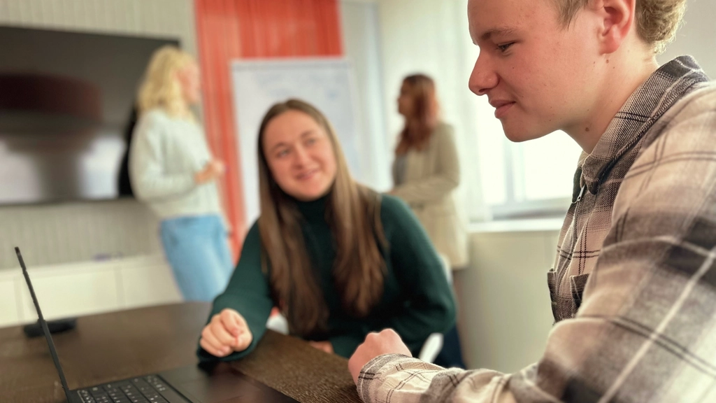 Zwei junge Menschen sitzen nebeneinander an einem Tisch in einem Besprechungsraum. Vor ihnen auf dem Tisch steht ein Laptop. Im Hintergrund sieht man unscharf zwei junge Frauen vor einem Whiteboard.