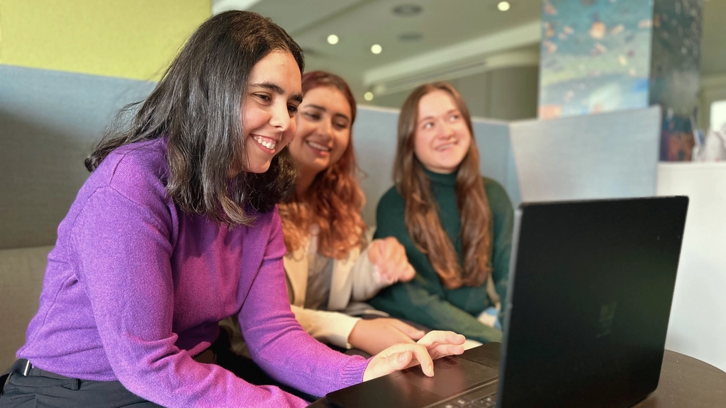 Drei junge Frauen sitzen auf einem Sofa nebeneinander. Vor ihnen auf einem Tisch steht ein Laptop.