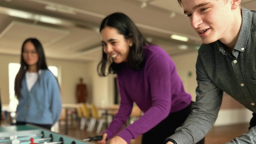 Zwei junge Menschen stehen nebeneinander an einem Kicker und spielen. Im Hintergrund ist eine weitere junge Frau zu erkennen.