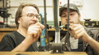 Ein jüngerer und ein mittelalter Mann stehen gemeinsam an einem Werktisch einer Fahrradwerkstatt und bedienen ein Werkzeug.