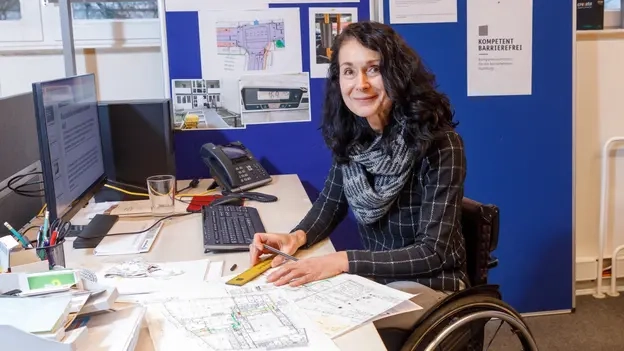 Eine Frau im Rollstuhl sitzt an einem Schreibtisch, hält ein Lineal und einen Bleistift in der Hand. Sie zeichnet auf Plänen.