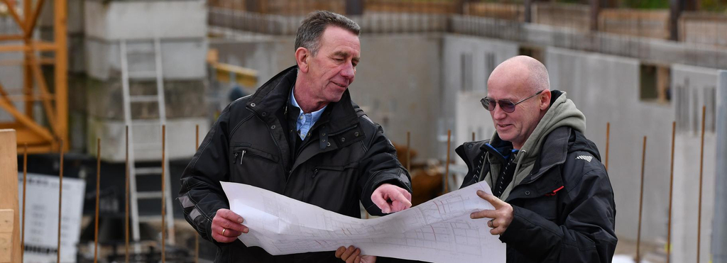 Zwei Männer stehen mit einem Bauplan in der Hand vor einer Baustelle, auf der ein Haus gebaut wird.