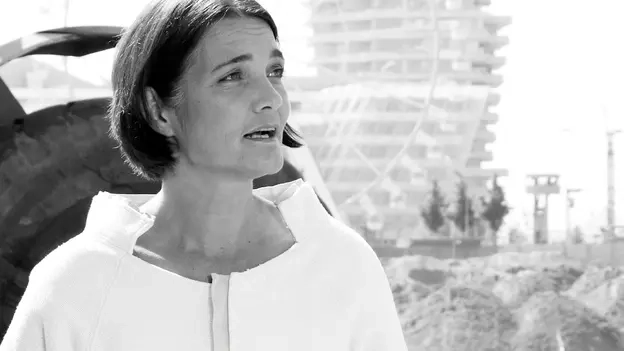 Schwarz-weiß Bild einer Frau im Rollstuhl auf einer Baustelle. Sie hält einen Gebäudeplan und einen Stift in der Hand.