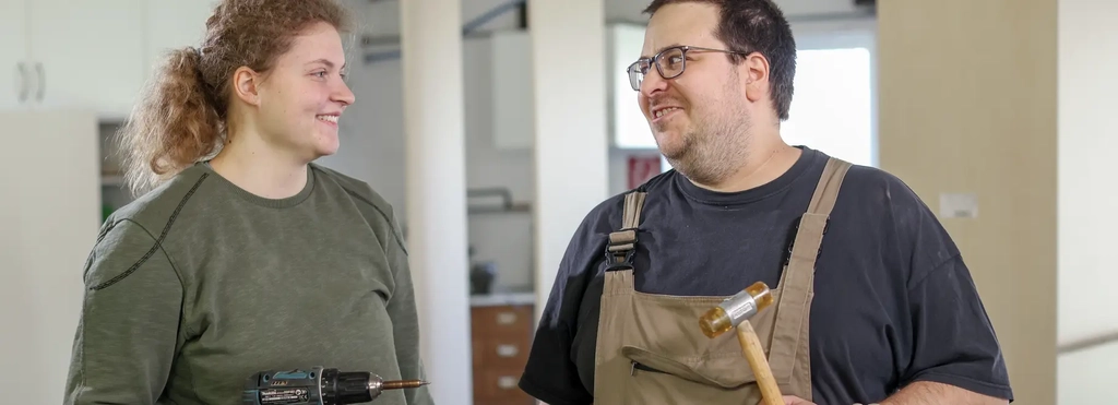 Eine Frau und ein Mann tragen Handwerker-Bekleidung und schauen sich lächelnd an. Beide haben Werkzeug in der Hand.