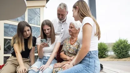 Drei junge Frauen, eine ältere Frau und ein älterer Mann sitzen auf einer Bank. Sie schauen alle auf ein Smartphone, das eine der jungen Frauen in der Hand hält.