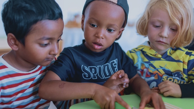 In einer Kita sitzen drei kleine Jungs zusammen am Tisch. Zwei von ihnen haben eine dunkle Hautfarbe und schwarze Haare, einer ist weiß und blond. Der Junge in der Mitte schneidet einen Apfel, die beiden anderen schauen konzentriert zu.