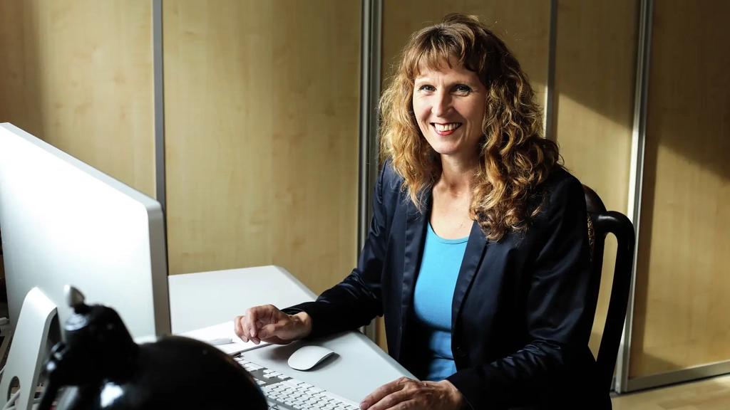 Eine Frau mittleren Alters mit langen dunkelblonden Locken sitzt an einem Schreibtisch. Vor ihr stehen eine Computermaus, eine Tastatur und ein Bildschirm. Sie trägt ein hellblaues T-Shirt und einen dunkelblauen Blazer.