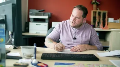 Ein Mann in einem lila T-Shirt sitzt an einem Schreibtisch im Büro. Er schaut auf den Computerbildschirm und schreibt etwas mit der Hand auf.