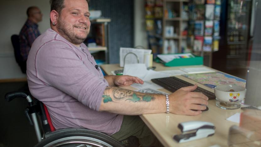 Ein Mann im Rollstuhl sitzt an einem Schreibtisch und lächelt in die Kamera.