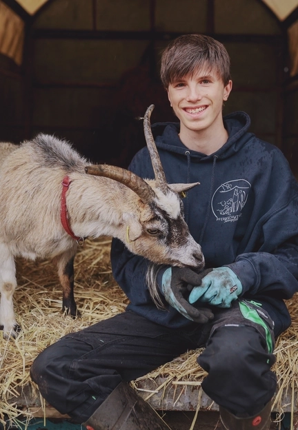 Ein junger Mann sitzt in einem Stall auf dem Boden. Eine Ziege neben ihm sucht nach Futter in seiner Hand.