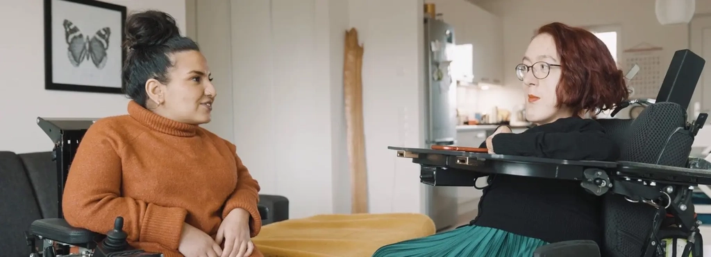 Zwei junge Frauen sitzen sich in ihren elektrischen Rollstühlen gegenüber und schauen sich an.