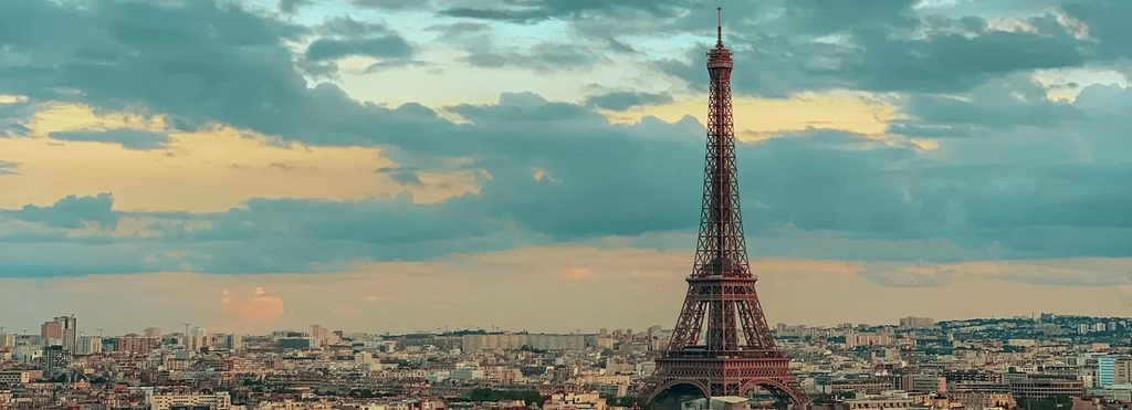 Eine Panoramaaufnahme von Paris aus der Luft. Hinten im Bild ist der Eifelturm zu sehen, der Himmel ist leicht wolkig und die Sonne geht unter.