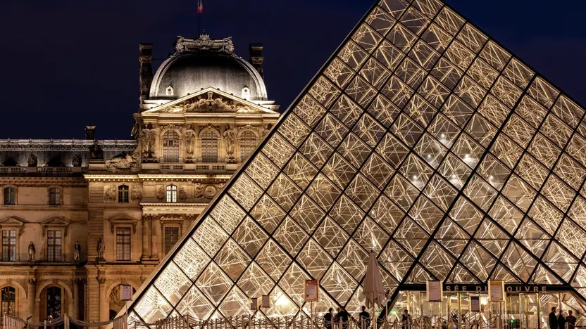 Zu sehen ist eine Nachtaufnahme vom Louvre in Paris. Vor dem Eingang warten Menschen