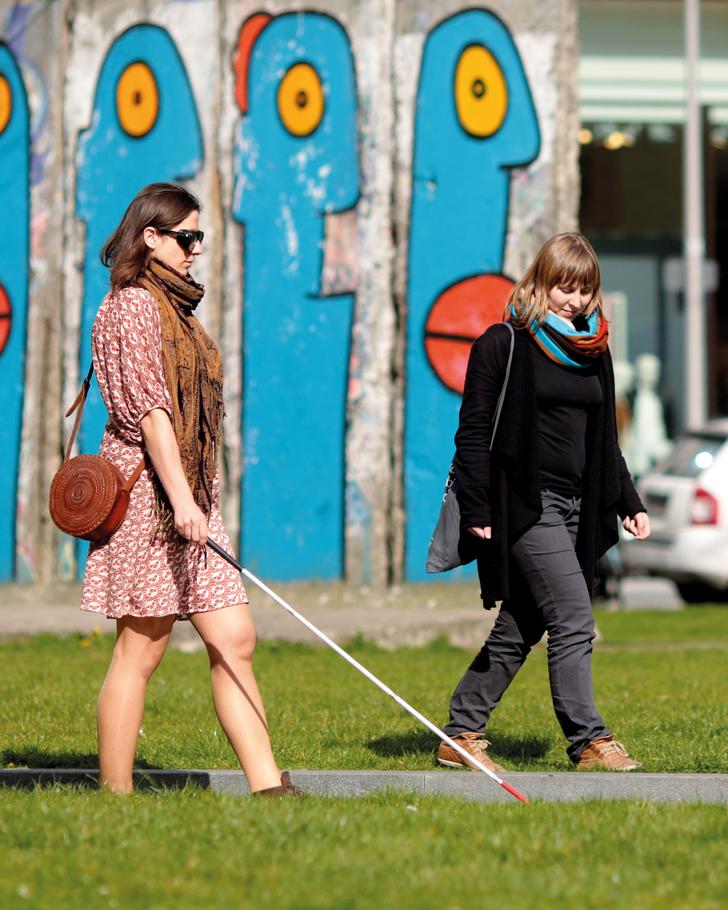 Zwei Frauen aus der Ferne fotografiert. Sie gehen einen Weg zwischen zwei Wiesen entlang. Eine von ihnen trägt eine Sonnenbrille. Sie nutzt einen Blindenstock.