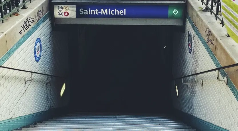 Zu sehen ist die Saint-Michel Bahnhaltestelle in Paris. Man blickt auf eine lange Treppe, die nach unten zur Haltestelle führt, darüber ist eine Fußgängerzone zu sehen