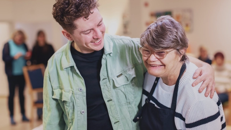 In einem Raum einer Begegnungsstätte: Zwei Menschen legen sich freundschaftlich die Arme um die Schultern und lachen. Der Linke ist ein junger Mann mit Jeansjacke und gewellten kurzen braunen Haaren. Die Rechte eine etwas kleinere, ältere Frau mit einer  Behinderung. Sie trägt eine Schürze über ihrem Pulli und hat eine Brille auf.