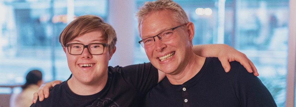 Ein Mann mit Down-Syndrom und ein Mann mit Brille haben einen Arm um die Schultern des anderen gelegt.