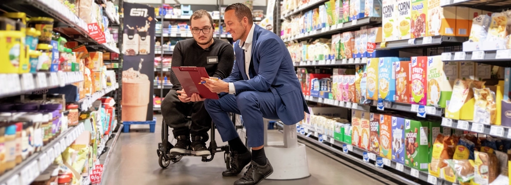 In einem Supermarkt-Gang zwischen zwei regalreihen sitzen zwei Männer. Links ein Mann in schwarzer Hose und Pulli im Rollstuhl mit Brille und mit Bart, rechts neben ihm ein Mann mit kurzen dunklen Haaren in blauem Anzug und weißem Hemd auf einem Tritthocker. Beide gucken in eine Liste.