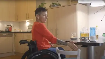 Eine Frau mit kurzen braunen Haaren und oranger Strickjacke sitzt in einem Rollstuhl in einer Küche. Vor ihr ist ein höhenverstellbarer Küchentisch zu sehen.