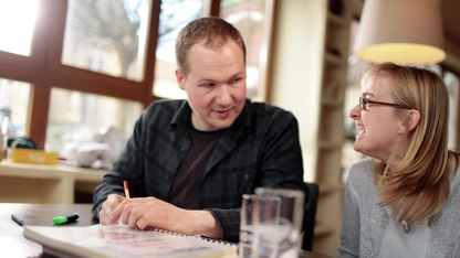 Ein Mann und eine Frau sitzen an einem Tisch und schauen sich an. Vor ihnen liegen Papiere und es stehen zwei Gläser auf dem Tisch.