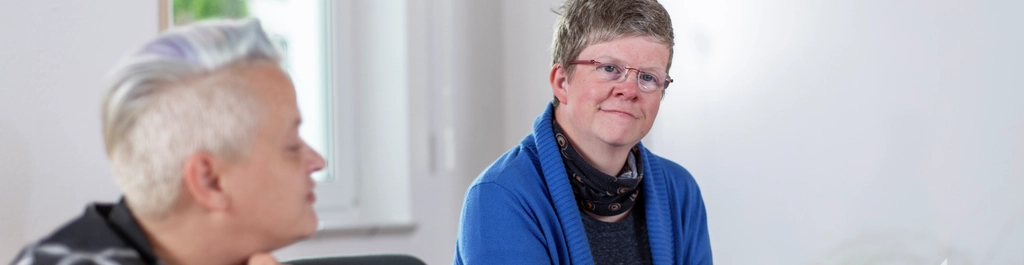 Zwei Frauen sitzen an einem Tisch und halten Papiere in ihren Händen. Die Umgebung sieht aus wie ein Büro.
