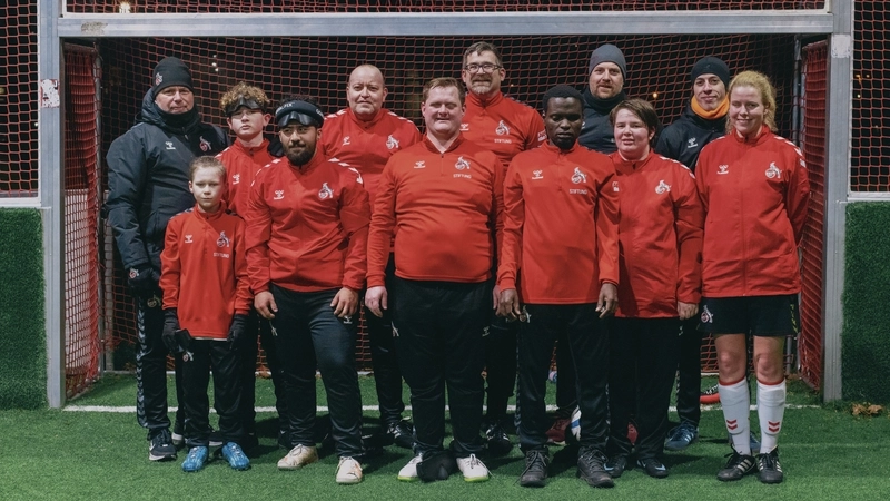 Gruppenfoto des Blinden-Fußball-Teams des 1. FC Köln. 9 Spieler*innen von jung bis alt und mit unterschiedlichen kulturellen Hintergründen stehen in roten Trainingsjacken in zwei Reihen hintereinander im Tor eines Kunstrasenplatzes. An den Rändern der hinteren Reihe stehen die drei sehenden Betreuer des Teams in schwarzen Trainingsjacken und mit schwarzen Mützen auf dem Kopf. Alle tragen das Vereinswappen des 1. FC Köln auf ihren Jacken.